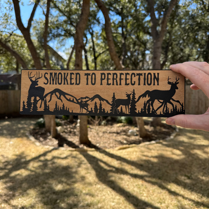 Hand holding a HipFlaskPlus Smoked to Perfection deer scene black and natural wood sign outdoors under oak trees