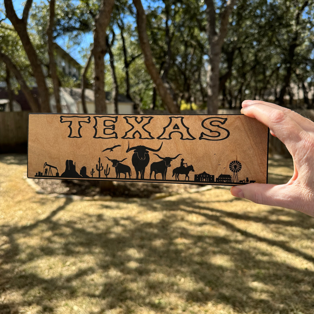 Hand holding a HipFlaskPlus Texas landscape black and natural wood sign outdoors under oak trees