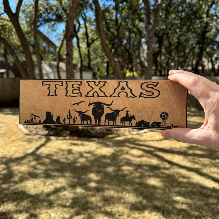 Hand holding a HipFlaskPlus Texas landscape black and natural wood sign outdoors under oak trees