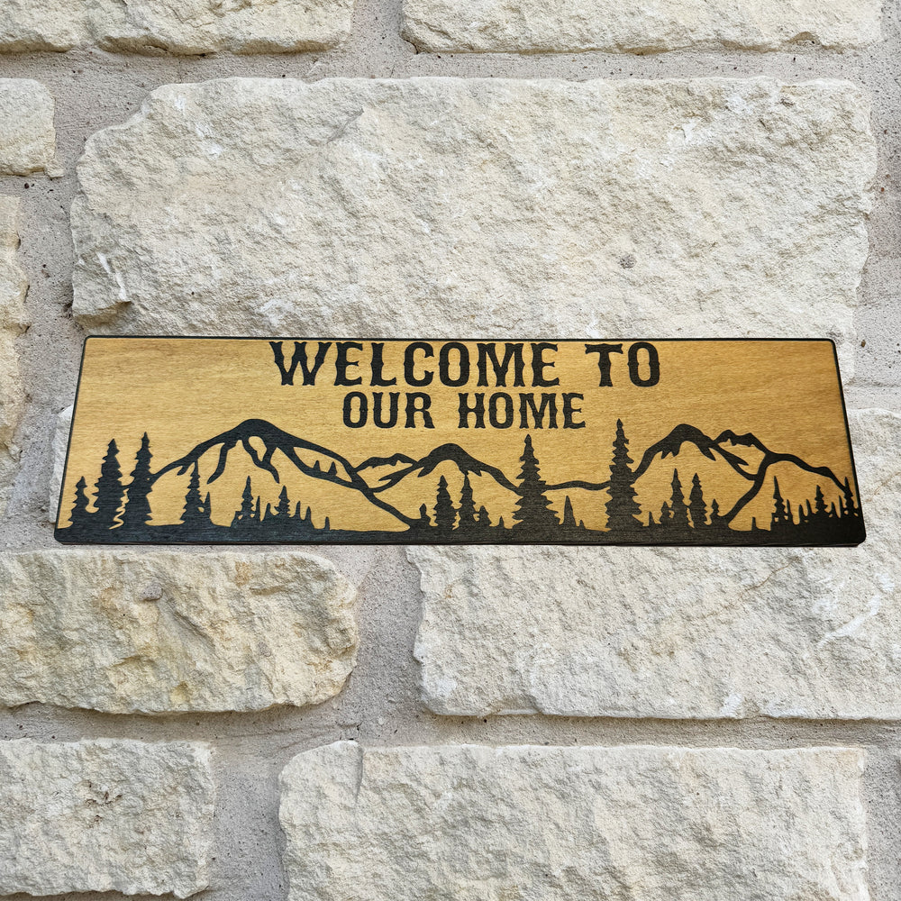 HipFlaskPlus Welcome to Our Home mountain scene black and natural wood sign on a stone wall