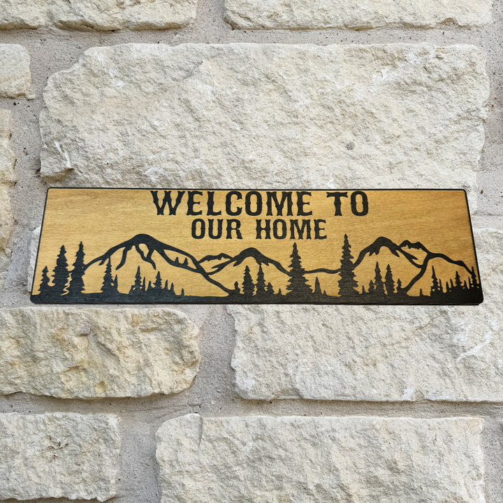 HipFlaskPlus Welcome to Our Home mountain scene black and natural wood sign on a stone wall
