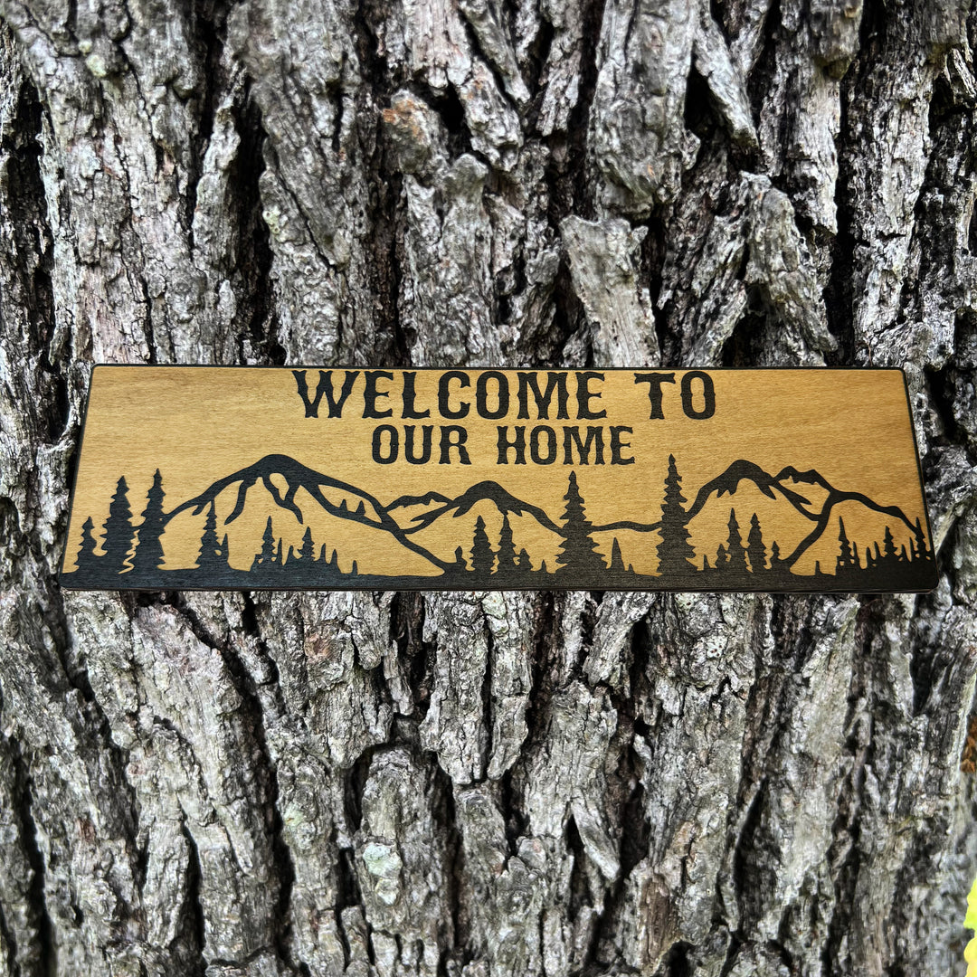 HipFlaskPlus Welcome to Our Home mountain scene black and natural wood sign on a tree trunk