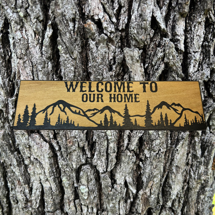 HipFlaskPlus Welcome to Our Home mountain scene black and natural wood sign on a tree trunk