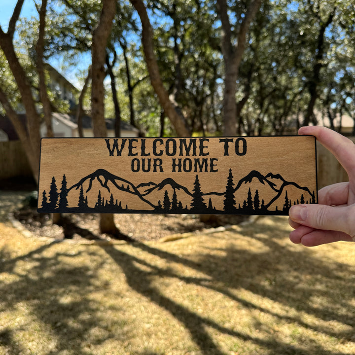 Hand holding a HipFlaskPlus Welcome to Our Home mountain scene black and natural wood sign outdoors under oak trees