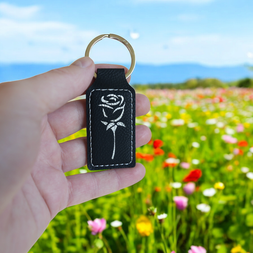HipFlaskPlus rectangular black leatherette keychain with a white rose engraving held against a blurred flower field