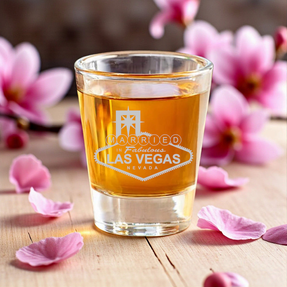 HipFlaskPlus 2oz clear glass shot glass with Married in Las Vegas engraving, set on a wooden table with pink flowers.