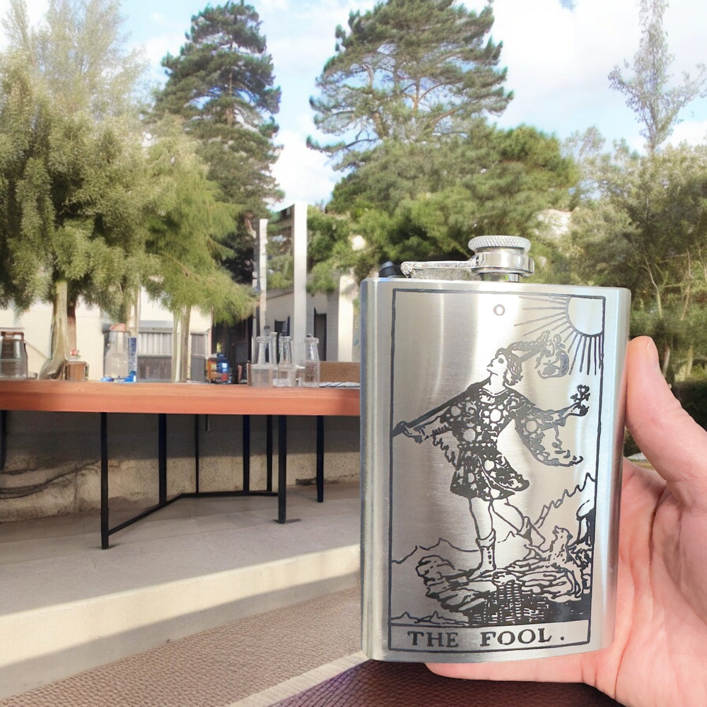 HipFlaskPlus 8oz stainless steel flask with laser engraved The Fool tarot card design held in a hand outdoors