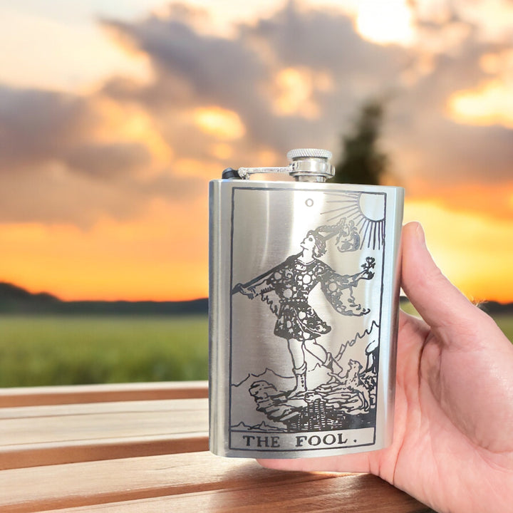 HipFlaskPlus 8oz stainless steel flask featuring a laser-engraved The Fool tarot card design held against a sunset.
