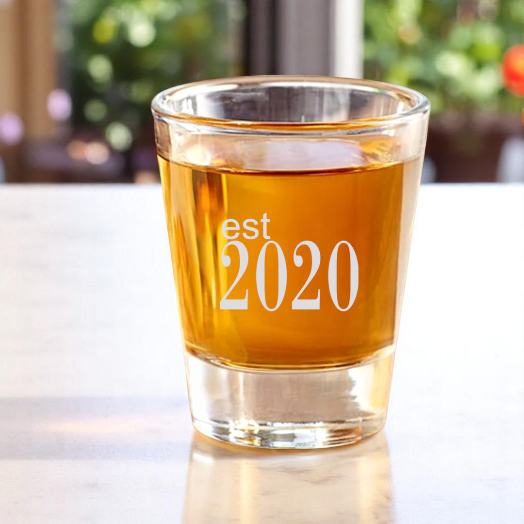 HipFlaskPlus 2oz shot glass filled with whiskey engraved with Est 2020 on a white table by a window