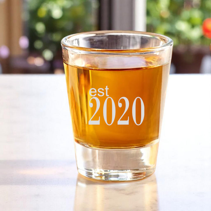 HipFlaskPlus 2oz shot glass filled with whiskey engraved with Est 2020 on a white table by a window