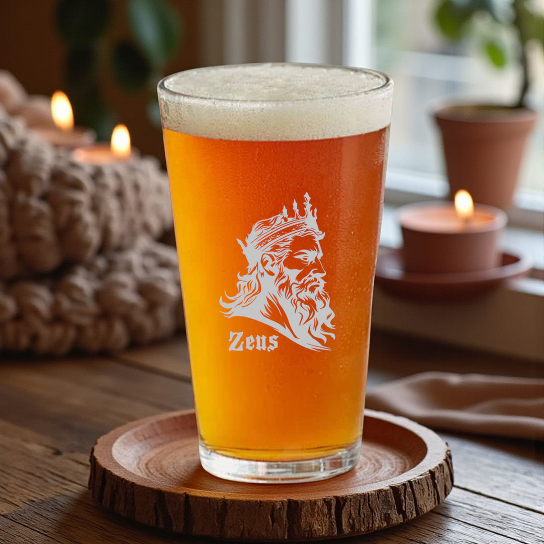 HipFlaskPlus 16oz pint beer glass filled with beer engraved with Zeus God of Thunder on a wood coaster beside a candle