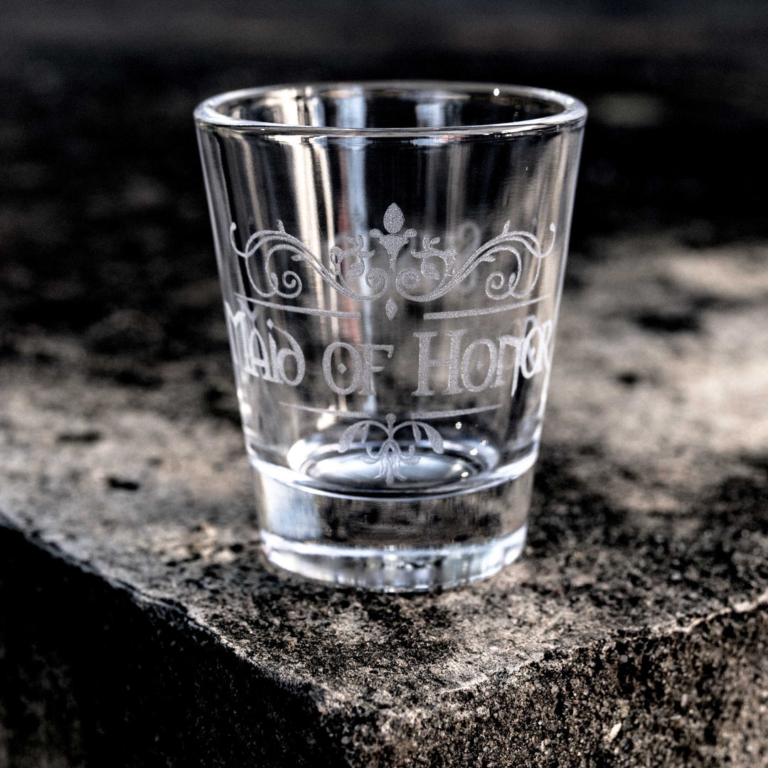 HipFlaskPlus 2oz clear shot glass engraved with a custom medieval Maid of Honor design on a stone ledge