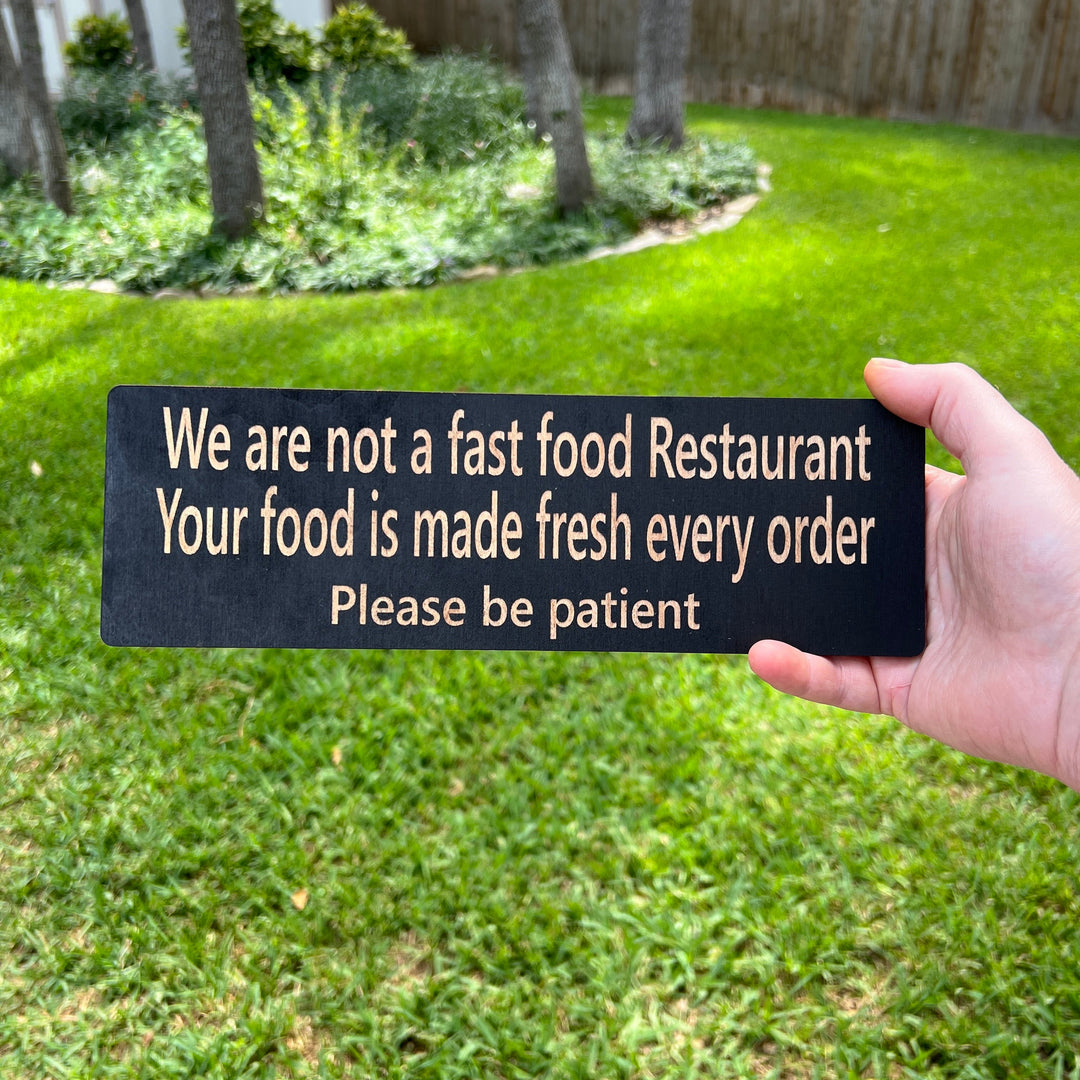 Hand holding a HipFlaskPlus custom We Are Not a Fast Food Restaurant Please Be Patient sign in black on a lawn