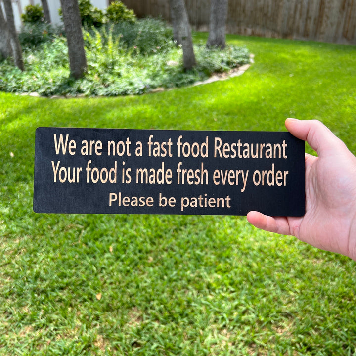 Hand holding a HipFlaskPlus custom We Are Not a Fast Food Restaurant Please Be Patient sign in black on a lawn