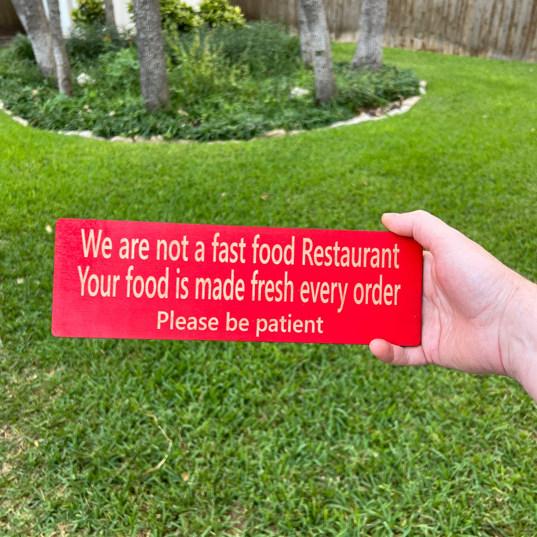 Hand holding a HipFlaskPlus custom We Are Not a Fast Food Restaurant Please Be Patient sign in red on a lawn